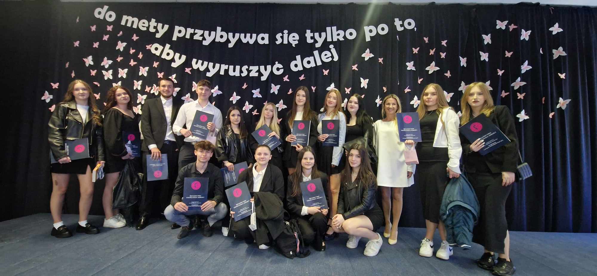 Grupa elegancko ubranych uczniów na scenie, w rękach trzymają świdectwa ukończenia szkoły, pomiędzy nimi stpi wychowawczyni w białęj sukience. W tle na granatowym tle zasłony biały napis "do mety przybywa się tylko po to, aby wyruszyć dalej".