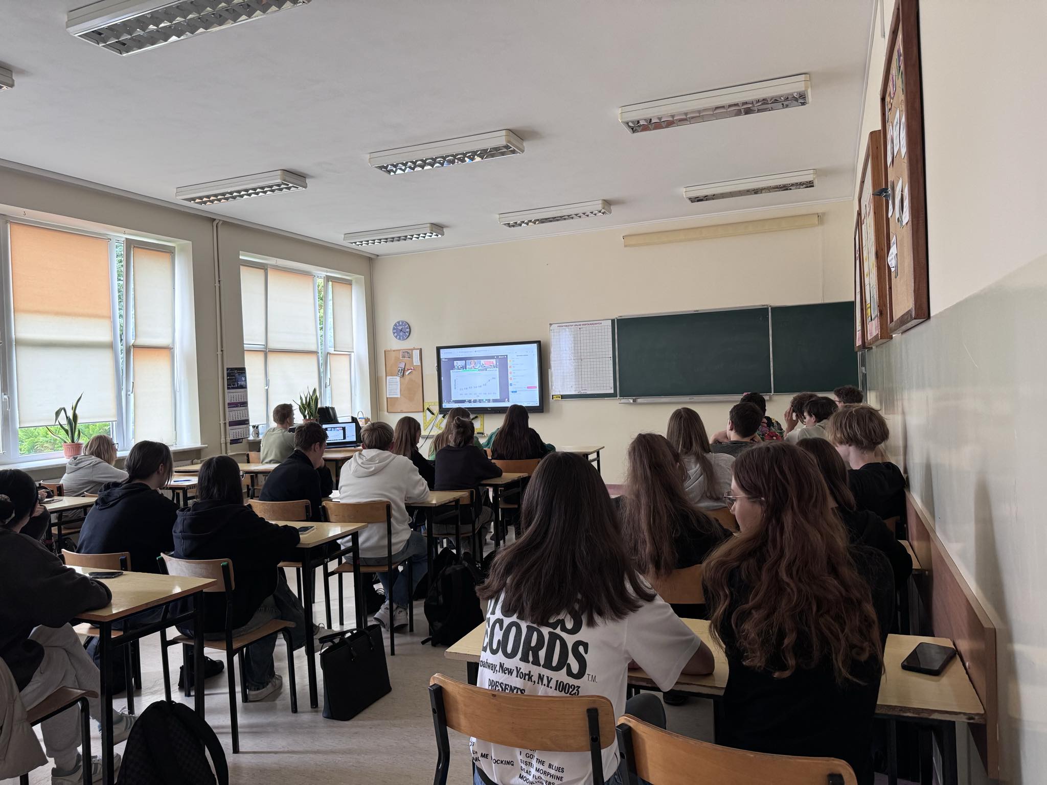 Ucznowie w sali lekcyjnej - widok zza pleców - siedzą w ławkach i patrzą w kierunku monitora.