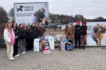 Grupa uczniów i nauczycieli stoi przez banerem Fundacji Kundelek, obok na chodniku stoją worki i puszki z karmą dla zwirząt oraz torby z akcesoriami.