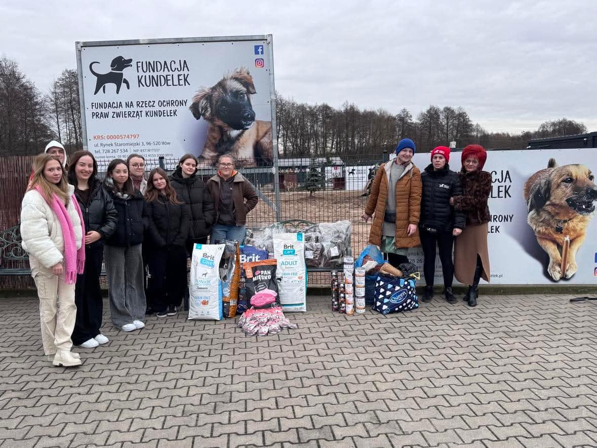 Grupa uczniów i nauczycieli stoi przez banerem Fundacji Kundelek, obok na chodniku stoją worki i puszki z karmą dla zwirząt oraz torby z akcesoriami.