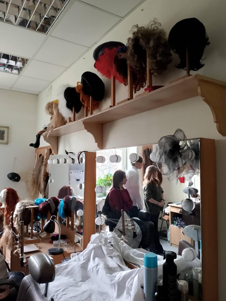 Charakteryzatornia teatralna. W lustrach odbicia zwiedzających ją uczniów. Na półce powyżej luster peruki i nakrycia głowy.