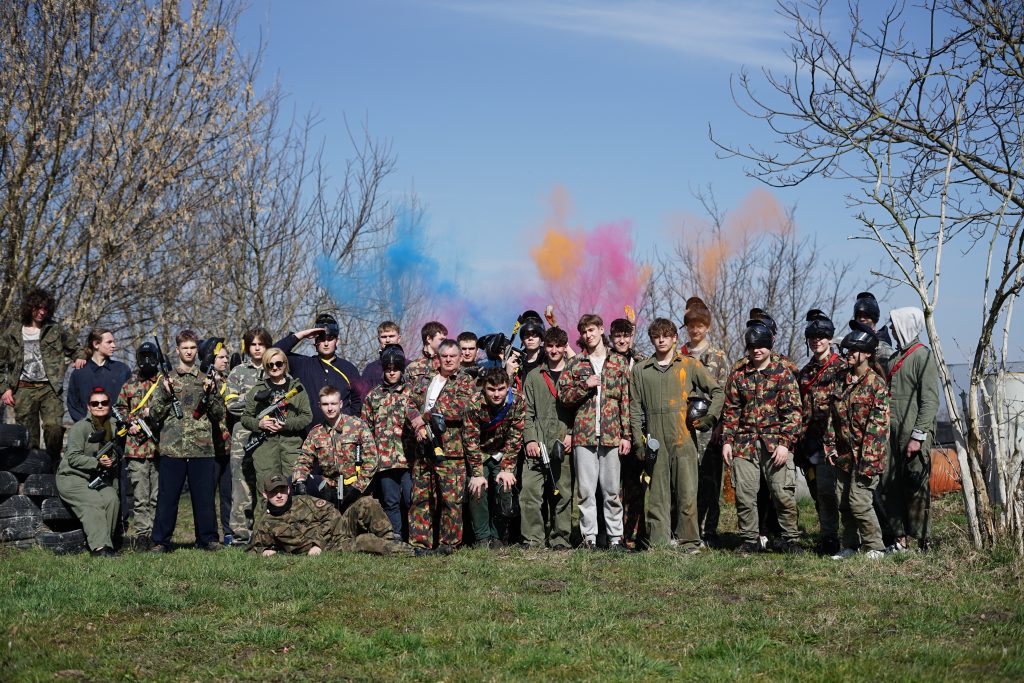 Duża grupa młodzieży wraz z nauczycielami w ochronnych strojach w barwach militarnych, z bronią paintballową w rękach na zielonej trawie.