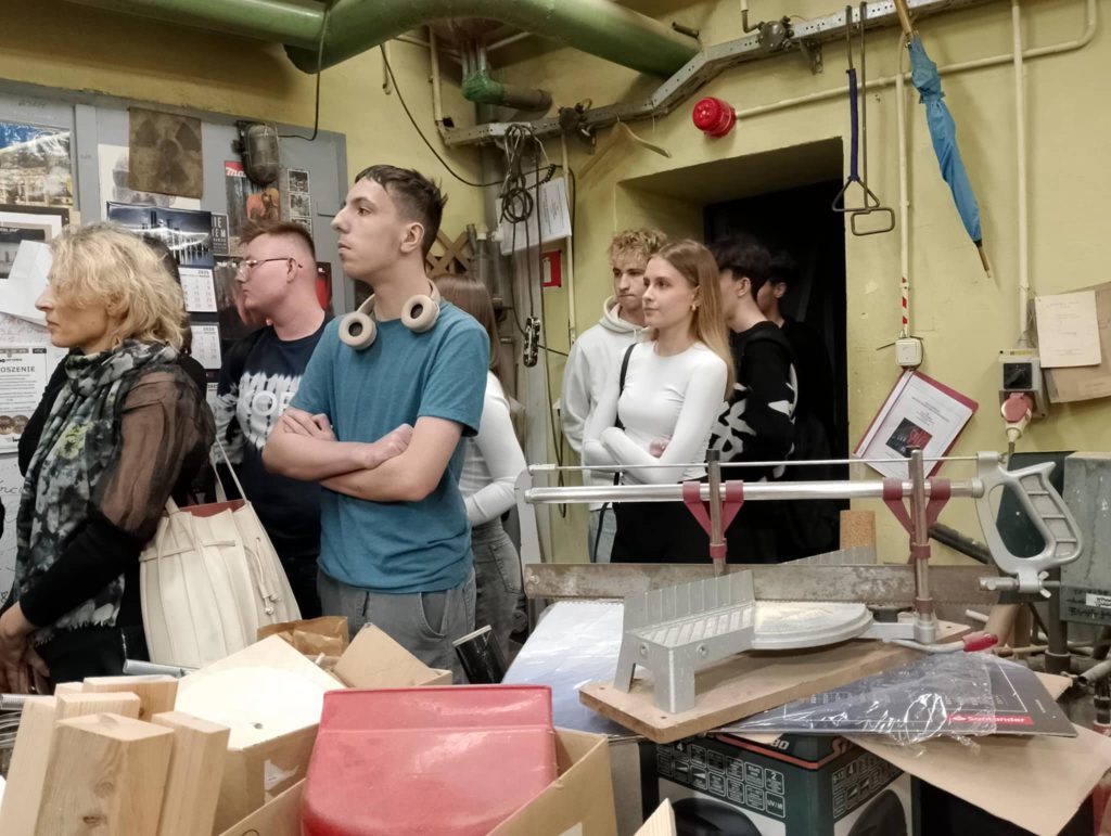 Teatralna pracownia stolarska. Uczniowie stoją wśród maszyn i urządzeń, obsewują z uwagą pracę rzemieślnika.